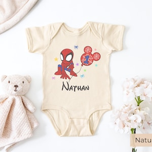 Natural color baby onesie with Spiderman birthday design, personalized name Nathan and age 1, styled with soft toy and floral decor
