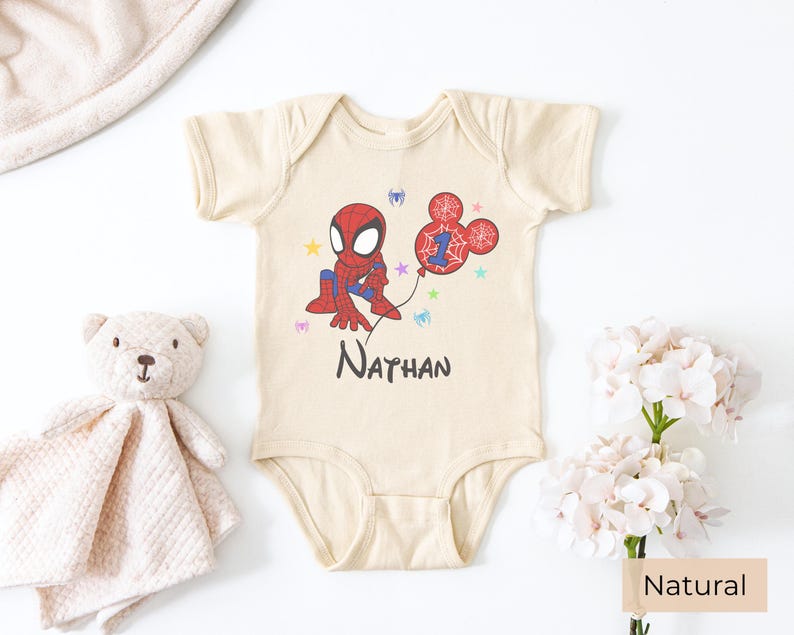 Natural color baby onesie with Spiderman birthday design, personalized name Nathan and age 1, styled with soft toy and floral decor