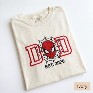 K&ouml;nnte beinhalten: Elfenbeinfarbenes T-Shirt mit rotem und schwarzem Stickdesign. Das Design zeigt das Wort "DAD" in roten Blockbuchstaben, eine Spider-Man-Maske und den Text "EST. 2026". Das Shirt ist gefaltet und hat ein Etikett mit der Aufschrift "Comfort Colors".