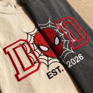 K&ouml;nnte beinhalten: Zwei T-Shirts, eines cremefarbenes und eines anthrazitfarbenes, mit einem Spider-Man-Motiv. Jedes Shirt zeigt den roten Buchstaben "D" mit roter Umrandung, eine Spider-Man-Maske und ein Spinnennetz-Design. Das cremefarbene Shirt hat "EST. 2026" in Wei&szlig;.