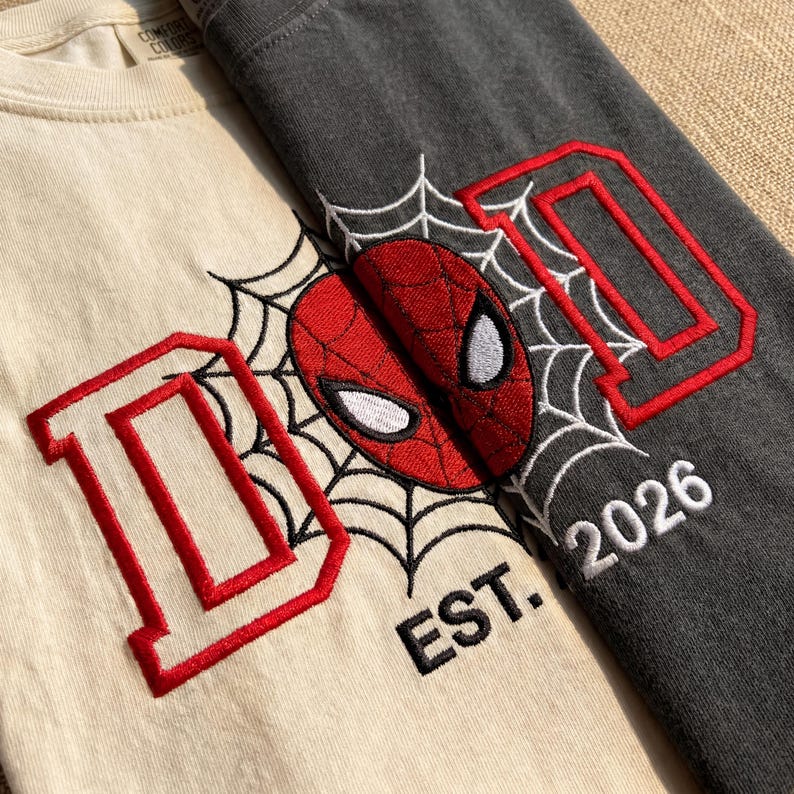 K&ouml;nnte beinhalten: Zwei T-Shirts, eines cremefarbenes und eines anthrazitfarbenes, mit einem Spider-Man-Motiv. Jedes Shirt zeigt den roten Buchstaben "D" mit roter Umrandung, eine Spider-Man-Maske und ein Spinnennetz-Design. Das cremefarbene Shirt hat "EST. 2026" in Wei&szlig;.