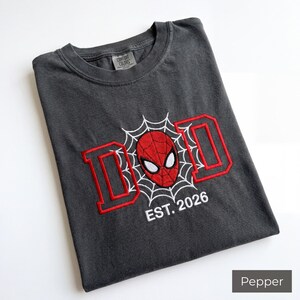 K&ouml;nnte beinhalten: Pfefferfarbenes T-Shirt mit rotem und wei&szlig;em Stickdesign. Das Design zeigt das Wort "DAD" in roten Blockbuchstaben, eine Spider-Man-Maske und den Text "EST. 2026". Das Shirt ist gefaltet und hat ein Comfort Colors-Etikett.