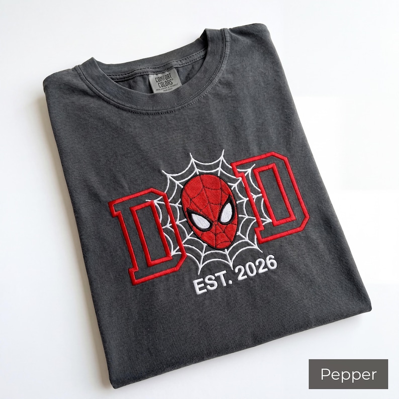 K&ouml;nnte beinhalten: Pfefferfarbenes T-Shirt mit rotem und wei&szlig;em Stickdesign. Das Design zeigt das Wort "DAD" in roten Blockbuchstaben, eine Spider-Man-Maske und den Text "EST. 2026". Das Shirt ist gefaltet und hat ein Comfort Colors-Etikett.