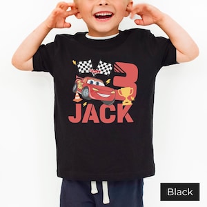 May include: A black short-sleeve t-shirt featuring a graphic of a red race car, a gold trophy, two checkered flags, and the number 3. The name JACK is printed in large red letters below the car. The design includes yellow lightning bolt accents.