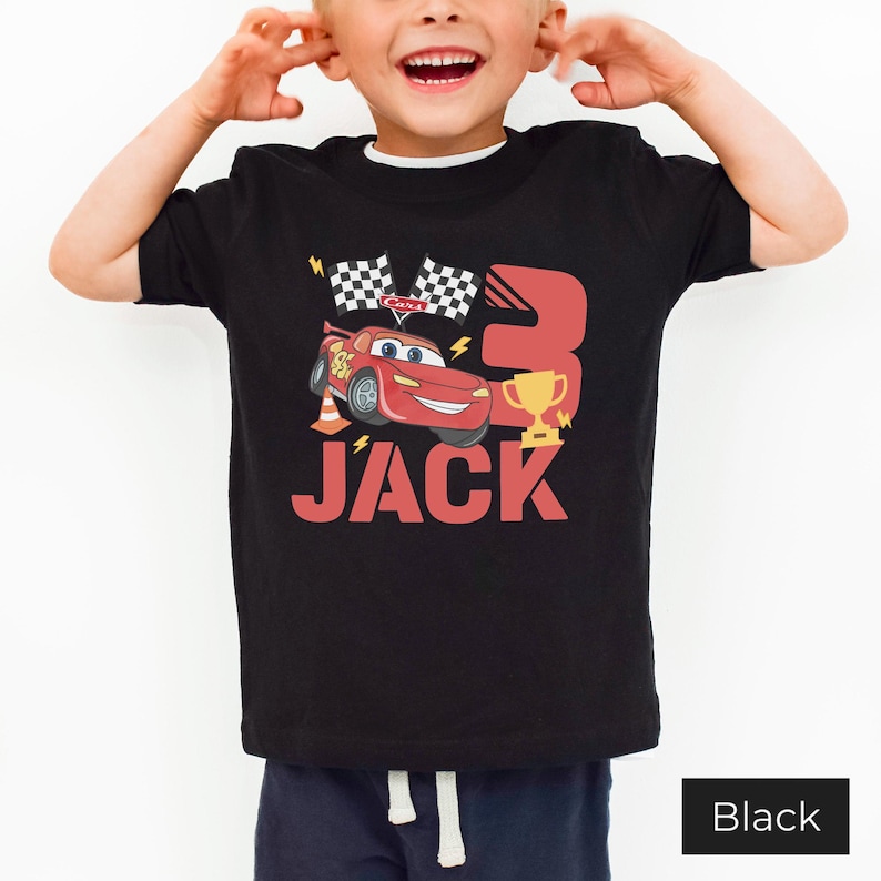 May include: A black short-sleeve t-shirt featuring a graphic of a red race car, a gold trophy, two checkered flags, and the number 3. The name JACK is printed in large red letters below the car. The design includes yellow lightning bolt accents.