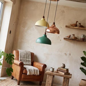 May include: Three pendant lights with wooden accents hang from the ceiling. One is yellow, one is copper, and the other is green. A brown leather armchair with a blanket sits below the lights. Wooden shelves hold decorative ducks and a ceramic pitcher.