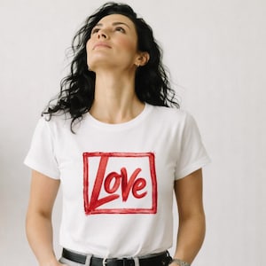 Könnte beinhalten: Weißes T-Shirt mit dem Wort "Love" in Rot in einem roten Quadrat. Das T-Shirt wird mit hellgrauen Jeans und einem schwarzen Gürtel getragen. Die Person, die das T-Shirt trägt, blickt nach oben. Das T-Shirt ist kurzärmlig.
