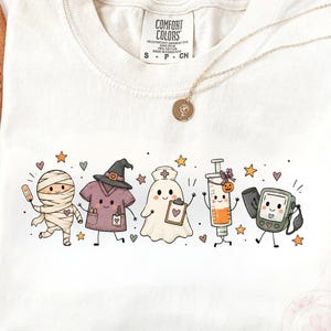 May include: A cream-colored t-shirt featuring a graphic print of five cartoon characters: a mummy, a scrub top with a witch hat, a ghost nurse with a clipboard, a syringe with a pumpkin, and a blood pressure monitor, all surrounded by small stars.
