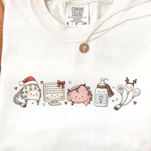 Può includere: Una t-shirt color crema con una stampa grafica raffigurante una cuffia EEG a cartone animato con cappello da Babbo Natale, un monitor con un fiocco rosso, un cervello con luci, una bottiglia etichettata EEG GEL con sciarpa e tre elettrodi.