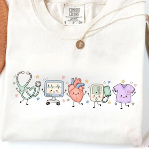 May include: A cream-coloured t-shirt featuring a graphic print of five anthropomorphic medical items: a green stethoscope, a heart rate monitor, a red heart, a blood pressure cuff, and a purple scrub top, all with smiling faces.