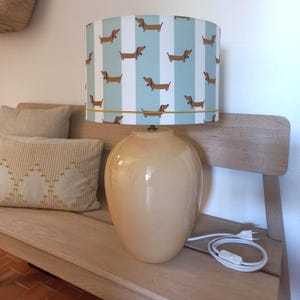 May include: A cream ceramic table lamp with a lampshade patterned with brown dachshunds on a light blue and white striped background. The lamp is on a wooden bench. The lamp has a UK plug.