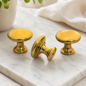Op de afbeelding: Drie messing kastknoppen met een gouden afwerking. De knoppen hebben een afgeronde bovenkant en een voet. Eén knop ligt op zijn kant, terwijl de andere twee rechtop staan. De knoppen liggen op een wit marmeren oppervlak.