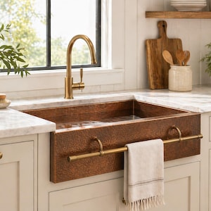 May include: A copper farmhouse sink with a brass faucet is installed in a kitchen. The sink has a rectangular basin and a towel bar. A white towel hangs from the bar. The kitchen has white cabinets and a marble countertop.
