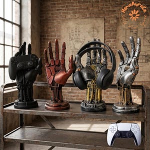 May include: Four robotic hand-shaped stands on a metal shelf. The first is matte black holding a game controller. The second is metallic copper. The third is weathered silver and gold holding black headphones. The fourth is silver. A white controller is below.