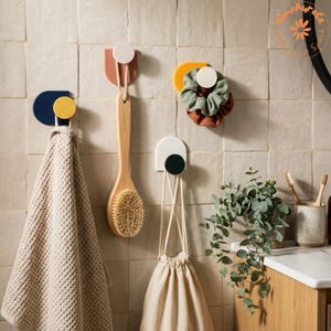 Könnte beinhalten: Vier Wandhaken mit geometrischen Formen in Marineblau, Terrakotta, Gelb und Weiß. Die Haken halten ein beiges Waffelpikee-Handtuch, eine Holzbadebürste mit Naturborsten, ein grünes und braunes Haargummi sowie einen beigen Beutel mit Kordelzug.