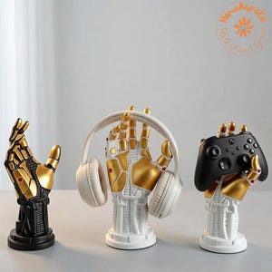 May include: Three robotic hand sculptures on circular bases. The left hand is black and gold. The center hand is white and gold, holding white over-ear headphones. The right hand is white and gold, holding a black wireless gaming controller.