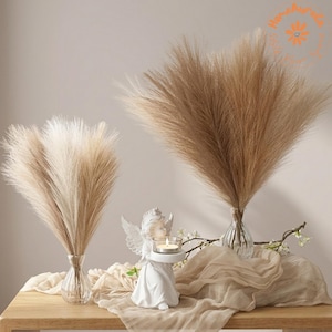 May include: Two clear glass vases containing fluffy, tan-colored pampas grass plumes. A white angel figurine holds a small glass candle holder with a lit tea light. The arrangement is set on a draped, light beige fabric cloth.