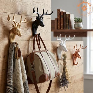 Può includere: Quattro ganci da parete a forma di testa di cervo con finiture oro, nero, bianco e rame. I ganci sono montati su un pannello in legno chiaro. Una sciarpa a motivi pende dal gancio dorato, una borsa da quello nero ed erbe da quello bianco.