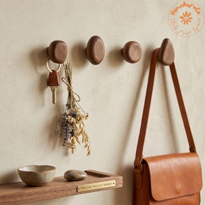 Puede incluir: Cuatro ganchos de pared redondeados de madera de nogal oscuro montados en una fila horizontal. Un gancho sostiene un llavero de metal con un ramo de flores secas. Un bolso de cuero marrón cuelga del gancho derecho. Debajo hay un estante de madera.