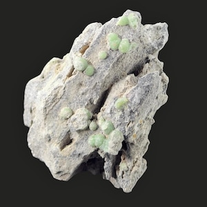 May include: A close-up of a rough, gray rock formation with several small, round, light green mineral deposits. The rock has a textured surface and is set against a dark background. The mineral deposits are clustered.