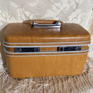 May include: A vintage tan suitcase with a brown handle and silver hardware. The rectangular case has a rounded top and bottom with a light gray trim. The suitcase has two silver latches and sits on small feet.