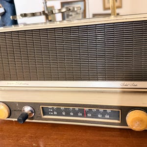 Può includere: Radio vintage Sears Silvertone a stato solido. La radio ha un corpo color crema con una griglia dell'altoparlante marrone scuro. È dotato di due manopole gialle e un quadrante di sintonizzazione con indicatori di frequenza.