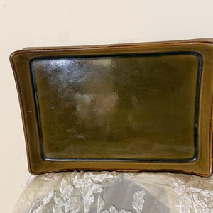 Puede incluir: Una bandeja rectangular de cerámica con interior verde oliva brillante y borde marrón oscuro. La bandeja tiene lados ligeramente curvos y un borde azul oscuro. La bandeja probablemente se utiliza para servir o exhibir.