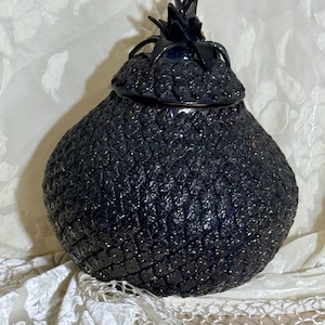 May include: A dark blue ceramic pineapple-shaped jar with a lid. The jar has a textured surface with a repeating pattern of small, raised shapes. The lid is topped with a spiky, dark blue crown.