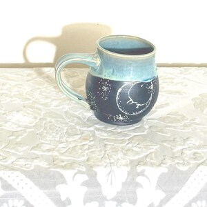 May include: A handmade ceramic mug with a blue and black glaze. The top half is a light blue, while the bottom half is dark blue with a white crescent moon and star design. The mug has a curved handle and is approximately 4 inches tall.
