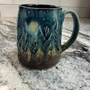 May include: A ceramic mug featuring a dark blue and teal glaze with a relief-carved forest scene. The design includes a pale moon, scattered stars, and textured pine trees that transition into a solid dark brown base at the bottom of the vessel.