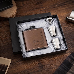 Puede incluir: Una petaca de metal forrada en cuero marrón con el texto Audrey, GROOMSMAN y 10.09.2026 grabado en el frente, presentada en una caja de regalo negra con dos vasos de metal plateado y un pequeño embudo de metal plateado sobre tela de satén blanca.