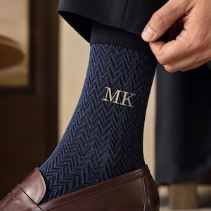 Op de afbeelding: Een marineblauwe sok met een donkerblauw visgraatpatroon en de geborduurde initialen MK in een lichtgrijs schreeflettertype. De sok wordt gedragen met een donkerbruine leren loafer en een donkere broek, reikend tot halverwege de kuit.