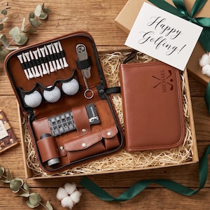 Könnte beinhalten: Ein Golf-Zubehörset aus braunem Leder in einer Geschenkbox. Das geöffnete Etui enthält weiße Golftees, drei Golfbälle, eine Pitchgabel aus Metall, einen Schlagzähler und eine kleine Tasche. Ein geschlossenes Etui ist mit dem Namen Michael personalisiert.