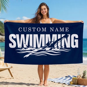 Op de afbeelding: Een rechthoekige marineblauwe strandhanddoek met de witte tekst CUSTOM NAME boven het woord SWIMMING in grote hoofdletters. Onder de tekst staat een witte grafische illustratie van een zwemmer die opspattend water veroorzaakt.