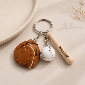 May include: A metal keychain featuring three charms: a brown leather baseball glove with visible stitching, a white baseball with red laces, and a light-colored wooden baseball bat engraved with the name WILLIAM in dark capital letters.