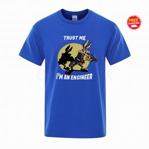 Peut inclure: T-shirt bleu roi avec un dessin animé de Wile E. Coyote tenant un appareil, avec le texte "TRUST ME I'M AN ENGINEER" en blanc. Le dessin est placé sur un cercle jaune. Un badge de livraison gratuite est dans le coin supérieur droit.