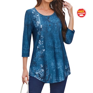 May include: A blue tunic top with a scoop neck and three-quarter sleeves. The top features a floral pattern on the front and three buttons. The fabric has a textured appearance. The model is wearing dark blue pants.