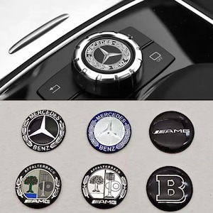 Op de afbeelding: Een verzameling Mercedes-Benz auto-emblemen en een bedieningsknop. De emblemen tonen het Mercedes-Benz logo, AMG en Brabus branding. De bedieningsknop is zilver en zwart met het Mercedes-Benz logo.