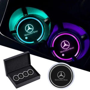 May include: Illuminated car cup holder inserts with the Mercedes-Benz logo. The inserts glow green and purple, and are shown in a car's cup holders. A black box contains four additional inserts. The inserts are black with a silver trim.