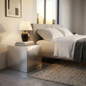 May include: A metallic silver cube side table with a smooth, reflective finish. A dark brown ceramic table lamp with a white tapered shade sits on top of the table, resting on a stack of two books. The table has a clean, geometric shape.