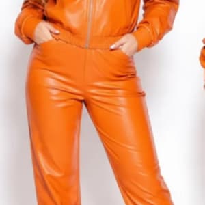 May include: Orange faux leather tracksuit featuring a zip-up hooded jacket and matching pants. The jacket has pockets and the pants have an elastic waistband. White sneakers complete the outfit.