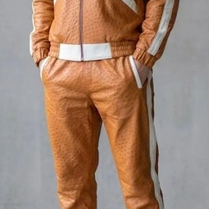 May include: A brown leather tracksuit with white detailing. The jacket features a zip-up front and a hood. White stripes run down the sides of the jacket sleeves and trouser legs. The trousers have an elasticated waistband. The person is wearing white trainers.