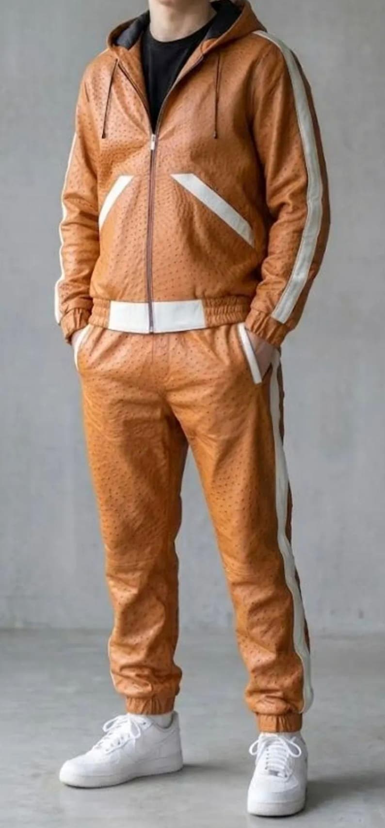 May include: A brown leather tracksuit with white detailing. The jacket features a zip-up front and a hood. White stripes run down the sides of the jacket sleeves and trouser legs. The trousers have an elasticated waistband. The person is wearing white trainers.