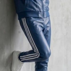 May include: A navy blue leather tracksuit with a zip-up jacket and matching trousers. The jacket and trousers have three white stripes. The outfit is completed with white trainers.