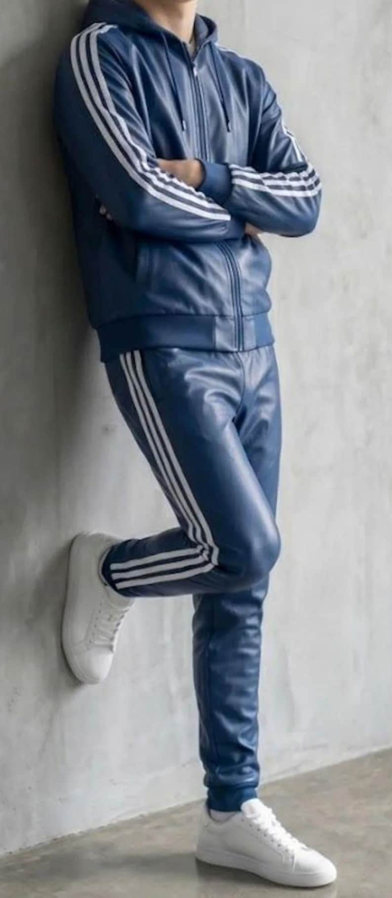 May include: A navy blue leather tracksuit with a zip-up jacket and matching trousers. The jacket and trousers have three white stripes. The outfit is completed with white trainers.