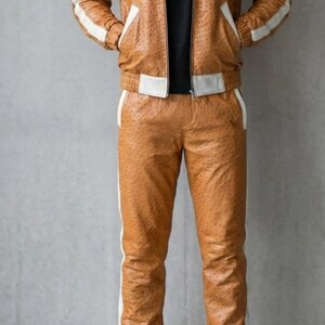 May include: A brown leather tracksuit with white stripes. The jacket has a hood and pockets, and the trousers have a matching stripe down the side. The outfit is paired with white trainers.