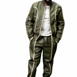 May include: Olive green track suit with cream-colored trim. The jacket has a zip-up front and the pants have a drawstring waist. The outfit is paired with white sneakers. The model is wearing a white t-shirt underneath the jacket.