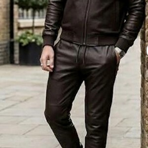 May include: A dark brown leather bomber jacket and matching leather trousers. The jacket features a zip closure and ribbed cuffs. The trousers have a drawstring waist. The outfit is completed with white trainers.