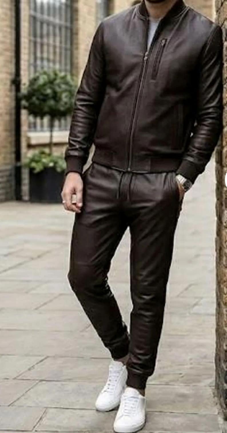 May include: A dark brown leather bomber jacket and matching leather trousers. The jacket features a zip closure and ribbed cuffs. The trousers have a drawstring waist. The outfit is completed with white trainers.