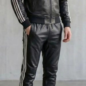 May include: Black leather tracksuit featuring white stripes on the jacket sleeves and trouser legs. The jacket is a bomber style with a zip closure. The trousers have a drawstring waist and the person is wearing white trainers.